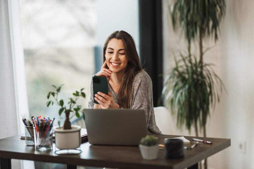How Much Does It Cost to Make a Landing Page? 2 shot-young-smiling-woman-using-cellphone-laptop-while-working-from-home