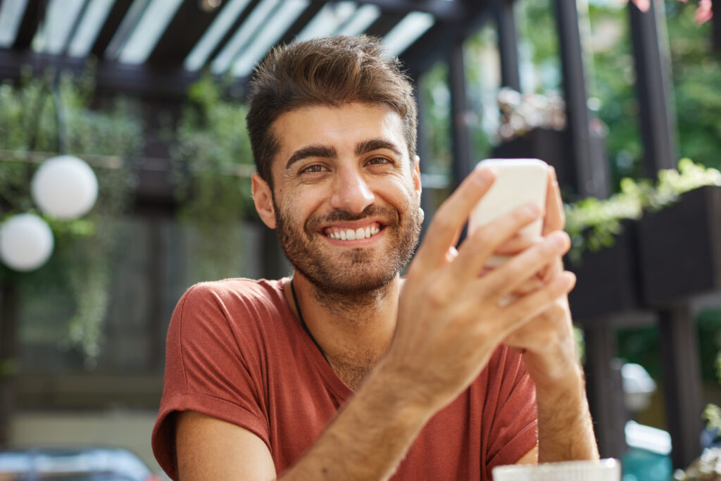 Outdoor shot of attractive man with smile and trendy hairstyle enjoying free wi-fi at summer platform of cafe, surfing internet on mobile phone, messaging friends online, inviting them to party.