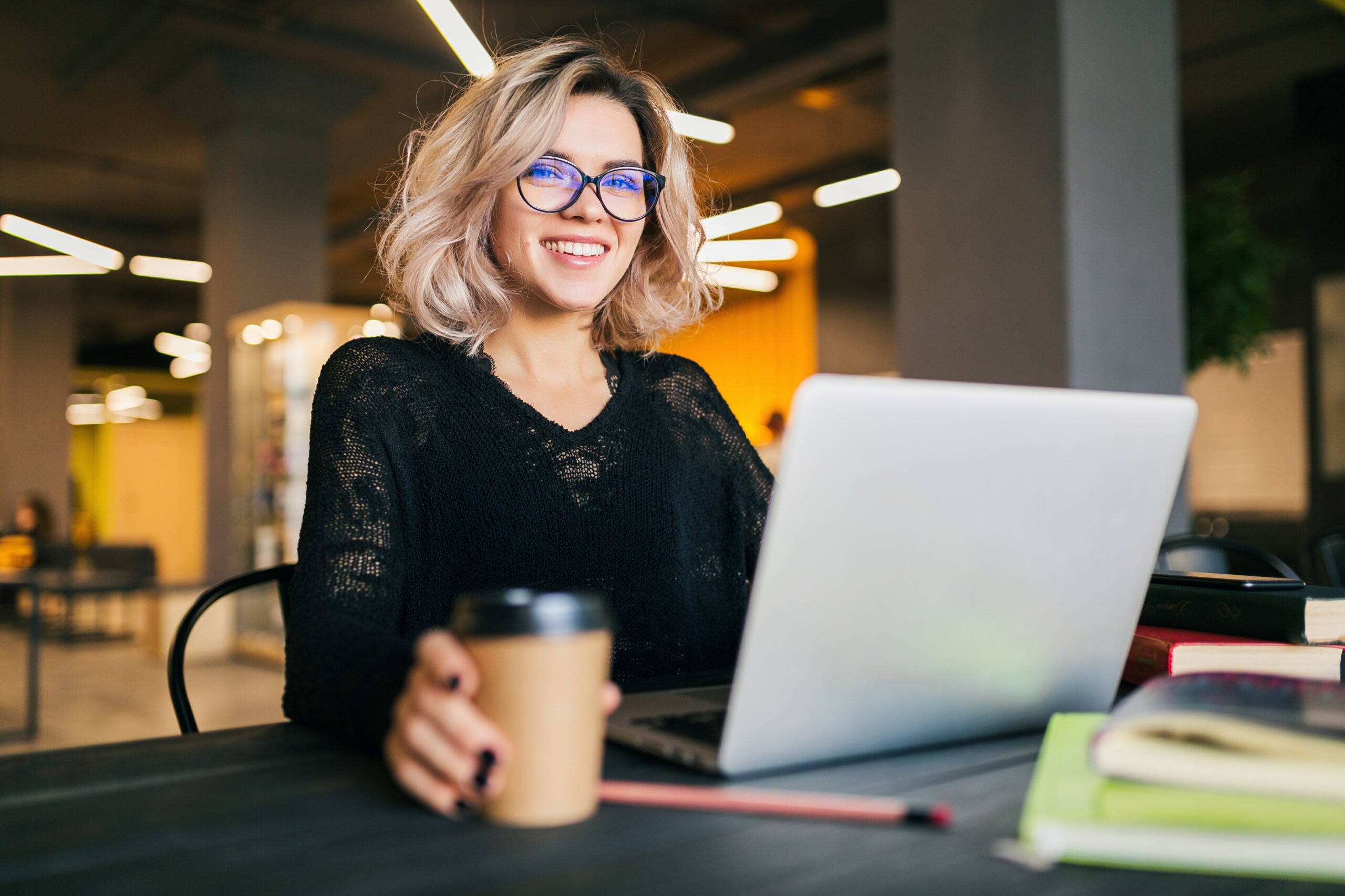 portrait of young pretty woman sitting at table in black shirt working on laptop in co-working office, wearing glasses, smiling, happy, positive, drinking coffee in paper cup