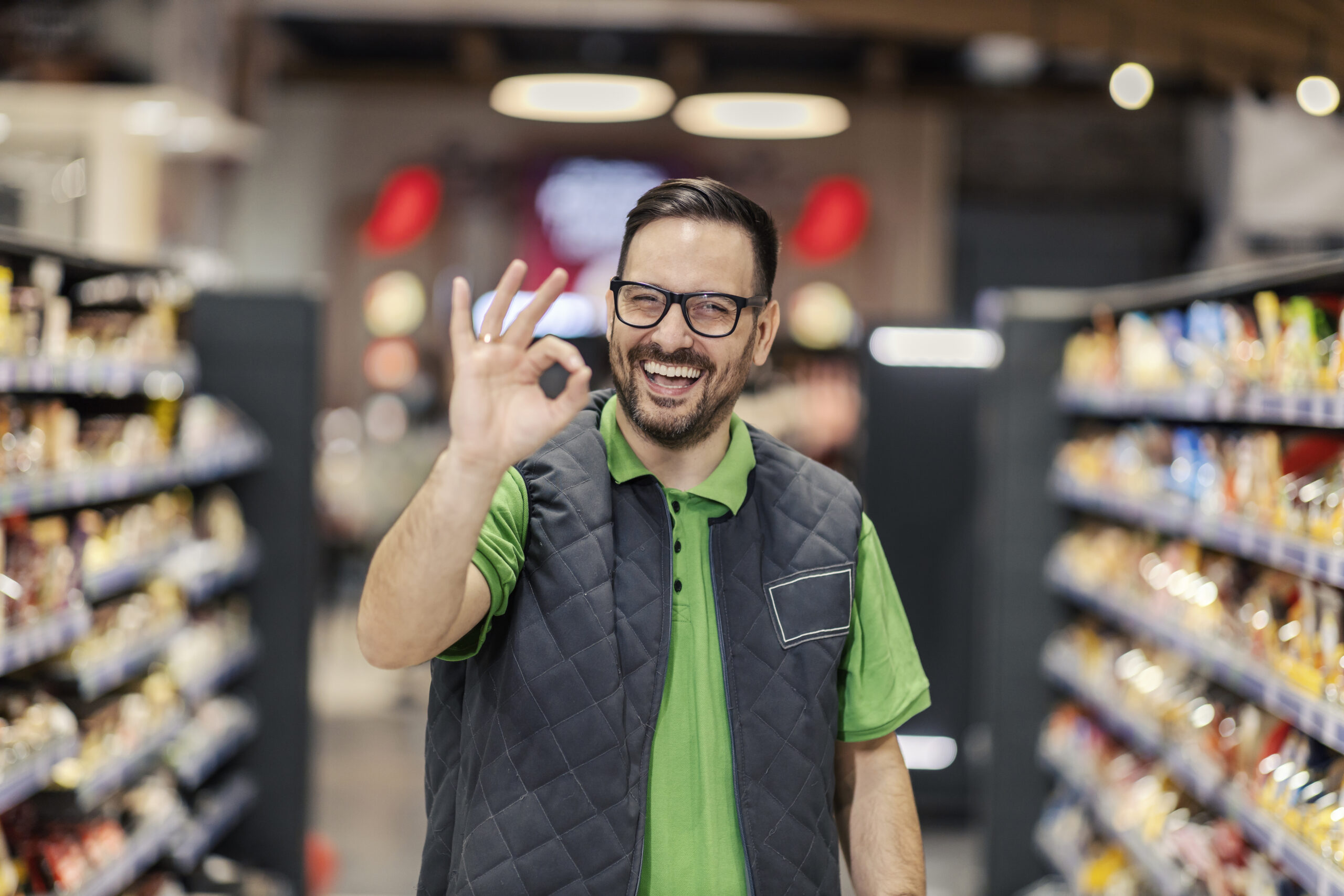 A salesman is standing at the marketplace and showing okay gesture at the camera.
