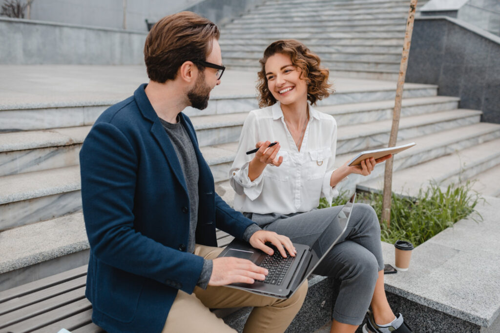 attractive-smiling-man-woman-talking-sitting-stairs-urban-city-center-making-notes